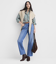 A woman in a green striped shirt, beige vest, and wide-leg jeans stands against a plain white background, holding a brown bag. carousel Product Image 3