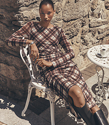 A woman in a plaid dress sits on a white chair in front of a stone wall, with a small table and coffee cup to her right. carousel Product Image 1