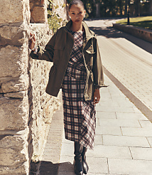 A woman in a plaid dress and olive jacket stands by a stone wall, with a paved path and trees in the background. carousel Product Image 1