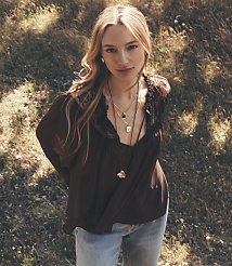 A young woman with blonde hair and multiple necklaces stands in the foreground, while a blurred background of dry grass and dappled sunlight creates a natural setting. carousel Product Image 1