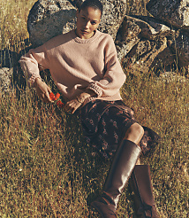 A woman in a pink sweater and floral skirt rests against rocks in a grassy field. carousel Product Image 1