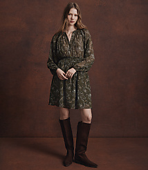 A woman in a patterned green dress and brown boots stands against a textured brown background. carousel Product Image 4