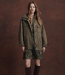 A woman in a green jacket and dress stands against a textured brown background. carousel Product Image 5