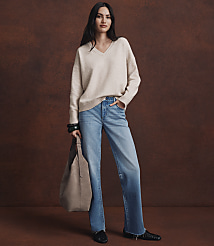 A woman in a cream v-neck sweater and light wash jeans stands against a textured brown background, holding a taupe tote bag. carousel Product Image 3