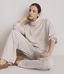 A woman in a beige loungewear set sits on a light-colored floor with a plain, light-colored wall behind her.