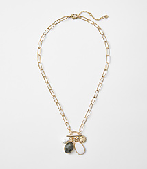 A gold paperclip chain necklace with a toggle clasp hangs against a plain white background, featuring three charms: a pearl, a dark oval stone, and a white teardrop-shaped stone. carousel Product Image 1