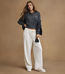 A young woman in a dark gray cardigan and white cable-knit pants stands against a plain brown background. carousel Product Image 4