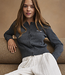 A young woman in a dark gray cardigan and white pants sits on a textured brown couch. carousel Product Image 4