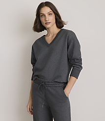 A woman in a dark grey v-neck sweatshirt and sweatpants stands against a plain grey background.