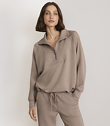 A woman in a taupe quarter-zip sweatshirt and matching sweatpants stands against a plain, light gray background.
