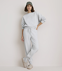 A woman in a grey sweatshirt and sweatpants stands against a plain, light-colored background, wearing a green baseball cap and slippers. carousel Product Image 2