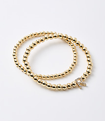 Two gold beaded bracelets, one with a diamond-encrusted "R" charm, are arranged on a plain white background. carousel Product Image 1
