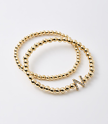 Two gold beaded bracelets, one with a diamond-encrusted letter "N" charm, lie on a plain white background. carousel Product Image 1