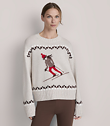 A woman in a cream-colored sweater with a skiing graphic stands against a plain, light gray background. carousel Product Image 2