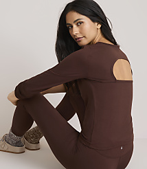 A woman in a brown long-sleeved shirt with an open back and matching leggings sits on a plain background, wearing fuzzy socks and slippers.