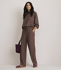 A woman in a brown ribbed loungewear set stands with a purple bag in front of a plain, light brown background.