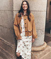 A smiling woman in a patterned dress and brown jacket leans against a stone pillar with a blurred building in the background. carousel Product Image 1