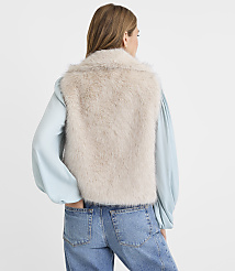 A woman in a light blue blouse and jeans stands with her back to the camera, wearing a fluffy cream-colored vest against a plain white background. carousel Product Image 3
