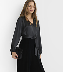 A woman in a black satin wrap top and velvet skirt holds a black clutch against a plain white background. carousel Product Image 2
