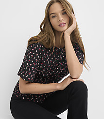 A young woman with long brown hair, wearing a black floral t-shirt and black pants, poses against a plain white background. carousel Product Image 1