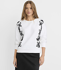 A woman in a white sweatshirt with black floral embroidery stands against a plain white background. carousel Product Image 1