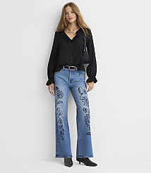 A woman in a black ruffled blouse and embroidered jeans stands against a plain white background. carousel Product Image 2