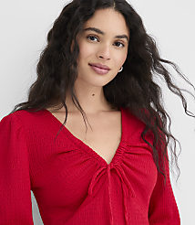 A woman with long, dark, wavy hair and a red, ruched V-neck top is shown against a plain white background. carousel Product Image 2