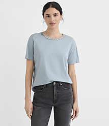 A woman in a light blue t-shirt with a decorative neckline and dark jeans stands against a plain white background. carousel Product Image 2