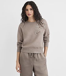A woman in a taupe sweater with floral embellishments stands against a plain, light gray background. carousel Product Image 2