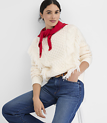 A woman in a cream cable-knit sweater with fringe and a red scarf around her neck poses against a plain white background. carousel Product Image 1