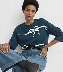 A woman in a teal sweater with white bow patterns and blue corduroy pants sits against a plain white background. carousel Product Image 1