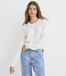 A woman in a cream-colored argyle sweater and light blue patterned jeans stands against a plain white background. carousel Product Image 1