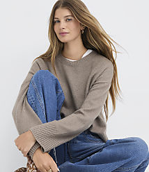 A young woman with long, blonde hair, wearing a brown sweater and blue jeans, sits against a plain white background. carousel Product Image 1