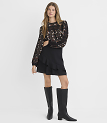 A woman in a floral blouse and black ruffled skirt stands against a plain white background. carousel Product Image 1