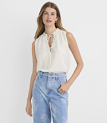 A woman in a cream-colored ruffled tie-neck blouse and light blue patterned jeans stands against a plain white background. carousel Product Image 2
