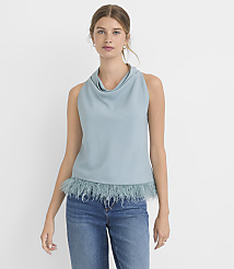 A woman in a blue cowl-neck sleeveless top with a feathered hem and blue jeans stands against a plain white background. carousel Product Image 1