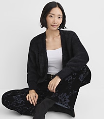 A woman wearing a black fuzzy cardigan, white top, and floral black pants sits against a plain white background. carousel Product Image 1