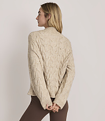 A woman wearing a beige cable-knit sweater and brown pants stands with her back to the camera against a plain light-colored background. carousel Product Image 3