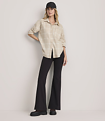 A woman wearing a beige plaid shirt, black flared trousers, and a black cap stands against a plain light-colored background. carousel Product Image 2