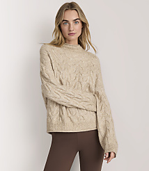 A woman wearing a beige cable-knit sweater and brown leggings stands against a plain white background. carousel Product Image 1