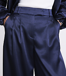 A person wears navy blue satin pleated trousers and a dark jacket over a white top against a plain light background. carousel Product Image 4