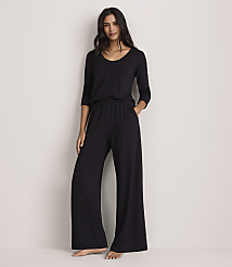 A woman wearing a black long-sleeved top and wide-leg black trousers stands against a plain, light-colored wall.