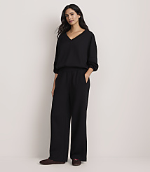 A woman wearing a black V-neck top and wide-leg black trousers stands against a plain, light-colored background.