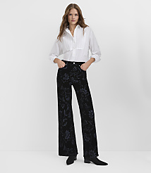A woman wearing a white pleated blouse and floral black velvet trousers stands against a plain white background. carousel Product Image 1