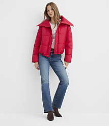 A woman wearing a red puffer jacket and blue jeans stands against a plain white background. carousel Product Image 2