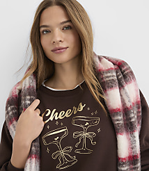 A woman wearing a brown "Cheers" sweatshirt and a red and white plaid scarf poses against a plain white background. carousel Product Image 1