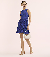 A woman wearing a royal blue lace dress and beige heels holds a white handbag against a plain light pink background. carousel Product Image 2