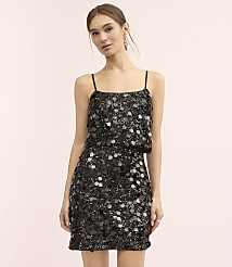 A woman wearing a black sequined mini dress stands against a plain light pink background.