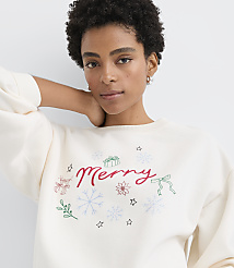 A woman with curly dark hair wearing a white "Merry" sweatshirt poses against a plain light gray background. carousel Product Image 1