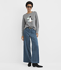 A woman wearing a grey penguin sweater, wide-leg blue pants, and black boots stands against a plain white background. carousel Product Image 3